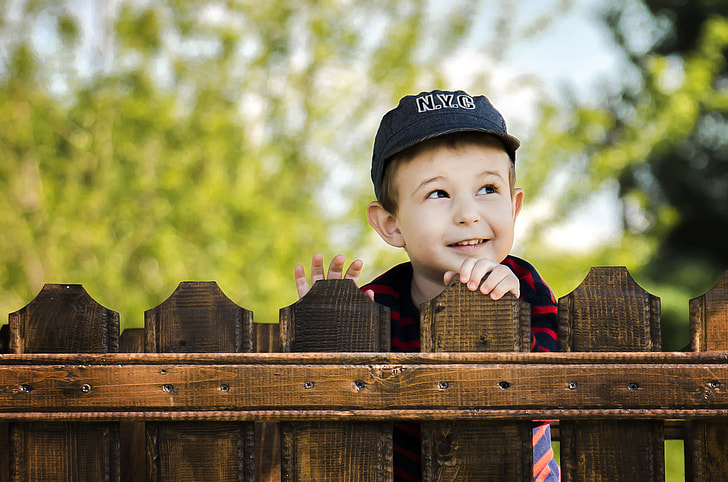 boy wearing black cap standing behind wooden fence