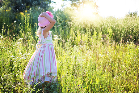 girl standing on green grass field while holding her pink hat during daytime