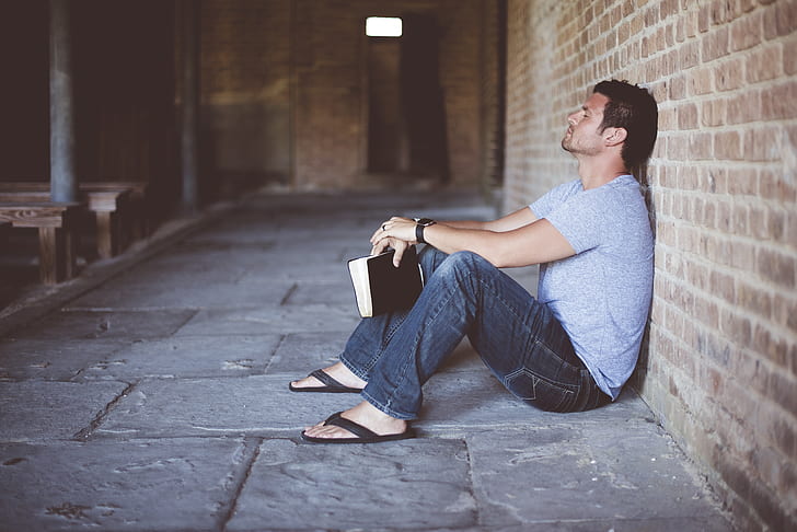 man wearing gray shirt holding book sitting beside brick wall