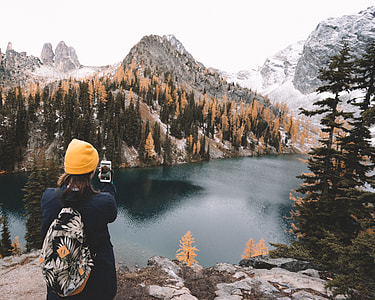 woman taking photo of lake near snow-covered mountain