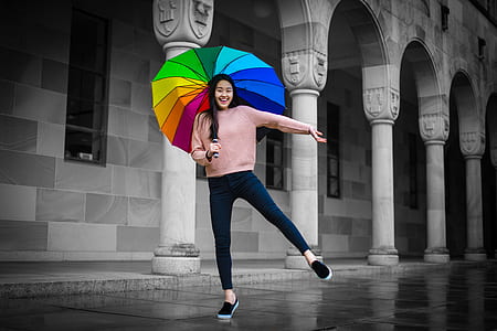 woman wearing pink crew-neck sweater, blue denim jeans and black slip-on sneakers holds multicolored umbrella