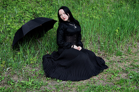 woman wearing black dress sitting on grass covered ground near umbrella during day