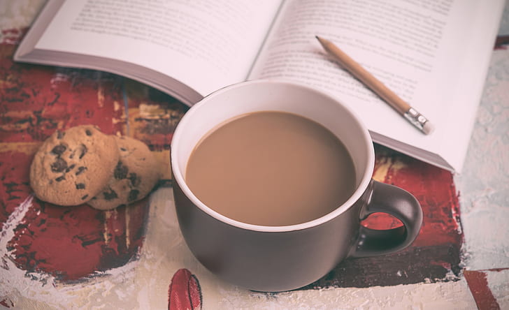 black mug filled with coffee beside open book and chocolate chip cookies
