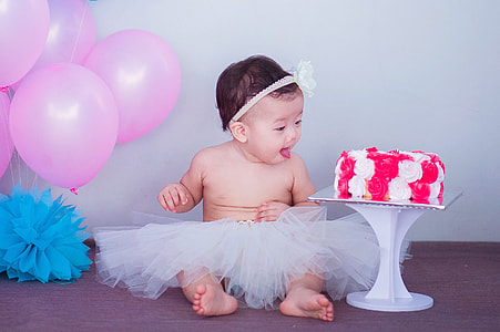 baby in white tutu skirt near white and red fondant cake and pink balloons