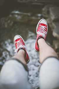 selective focus photography of man's feet wearing red lace-up low-top sneakers above body of water
