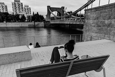 woman sitting on bench while reading book in front of body of water grayscale photo