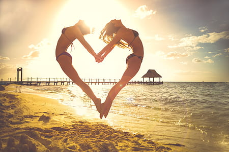 two women jumping while forming heart shape beside seashore during daytime