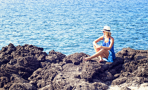 woman in blue scoop-neck sleeveless dress sitting on black rock near ocean at daytime