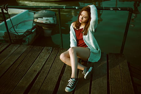 woman wearing red shirt, gray cardigan, and pair of black low-top sneakers outfit sitting on brown wooden dock