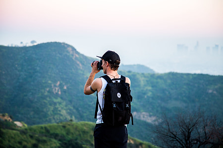 man stands on peak while taking picture of mountain at daytime