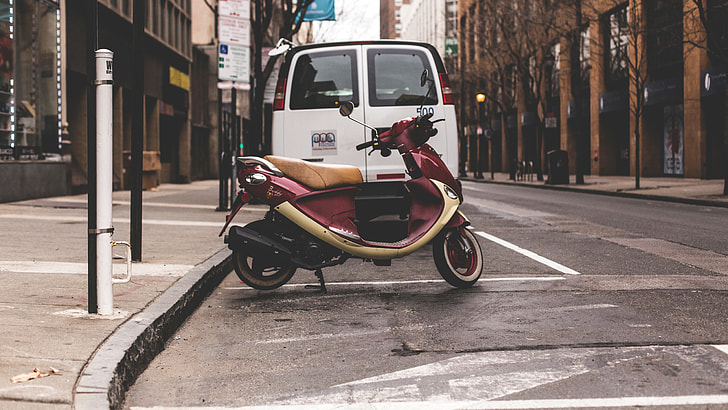 photography of red motor scooter near white van and brown concrete buildings