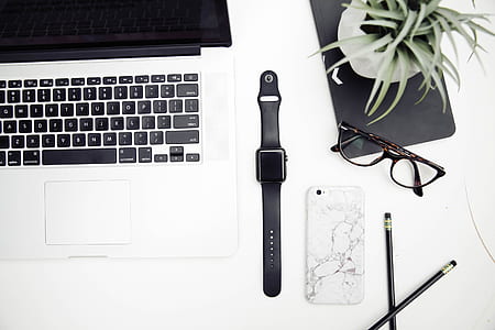 space gray aluminum case Apple Watch with black sports band near black frame wayfarer style eyeglasses and MacBook Pro