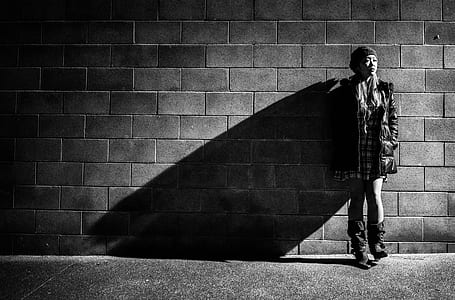 grayscale photography of woman leaning on brick wall wearing jacket and booties