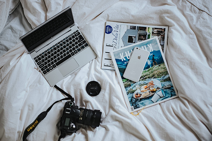 Silver laptop, a camera, magazines and other items on white bed sheets
