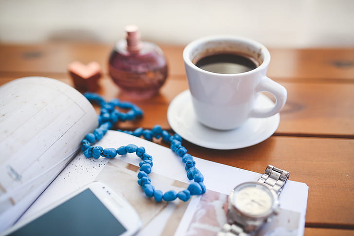 beaded blue necklace on book between white cup with coffee and analog watch