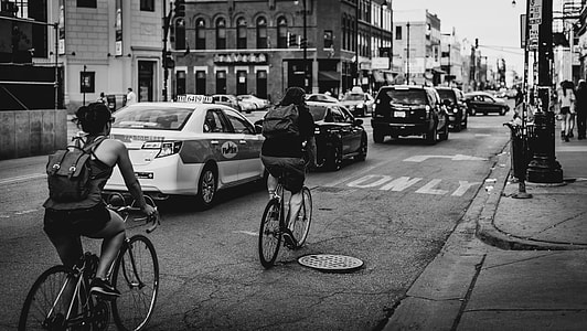 greyscale photo of people in bike riding along cars on road with buildings