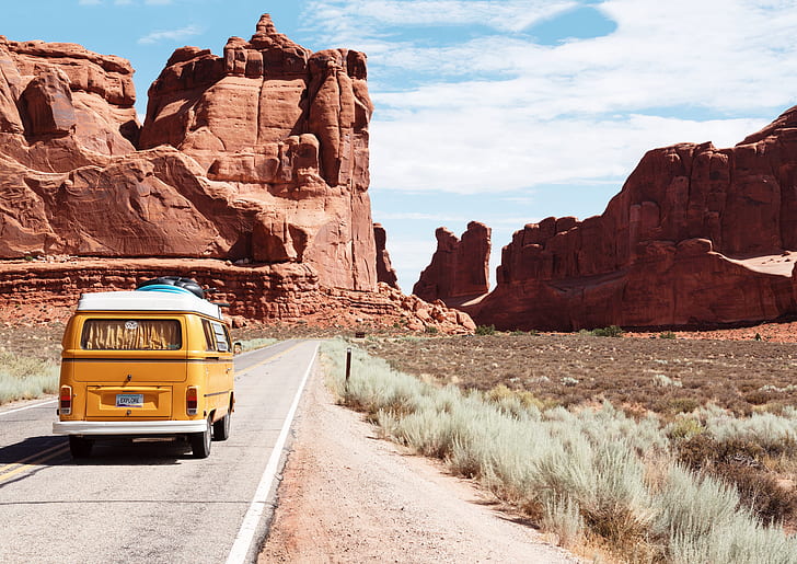 yellow van traveling on concrete road towards red rock monoliths during daytime