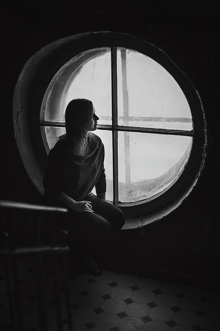 grayscale photo of woman in shirt standing beside round-shaped window
