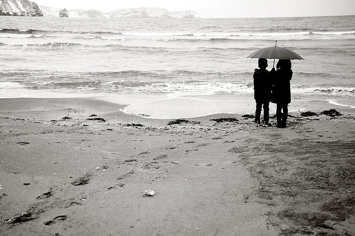 gray scale photo of two girls holding gray umbrella standing beside body of water