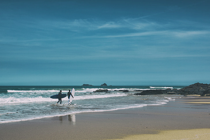 Two surfers walk along the beach on a sunny day on the coast of Cornwall in England