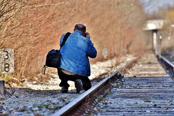 man wearing blue denim jacket and black pants kneeling beside train rail holding camera during daytime