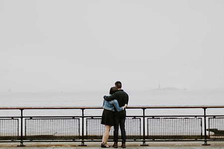 man and woman hugging beside black concrete fence in front of body of water during daytime