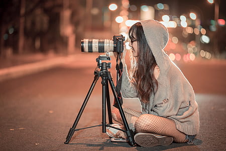 woman in grey hooded jacket sits on road using DSLR camera with tripod