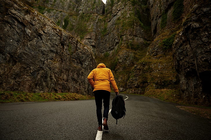 photo of person wearing yellow hoodie and black pants holding backpack walking on roadway between mountain