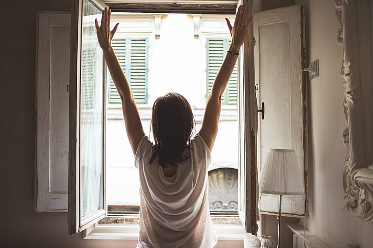 woman standing in front of opened window and putting her hands in the air