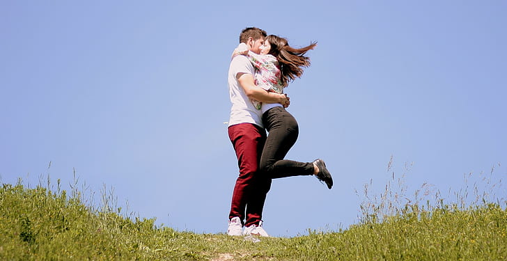 girl hugging boy with raising left foot on air surrounded by green grass in the hill under blue sky during daytime
