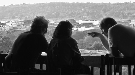 grayscale photo of three people leaning on wood rail talking
