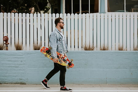 man in blue denim button-up jacket and black pants holding multicolored longboard during daytime