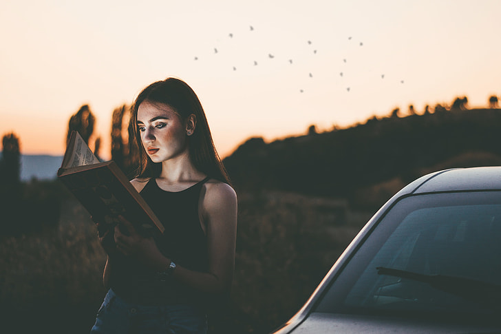 photo of a woman reading book while leaning on car during daytimne