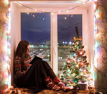 woman in brown and black sweater and black bottoms with socks sitting besides white wooden framed clear glass window pane with string light in front of mini Christmas tree reading book