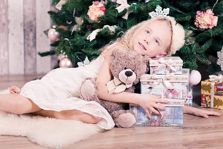 child lying on floor near christmas tree hugging teddy bear and gift boxes