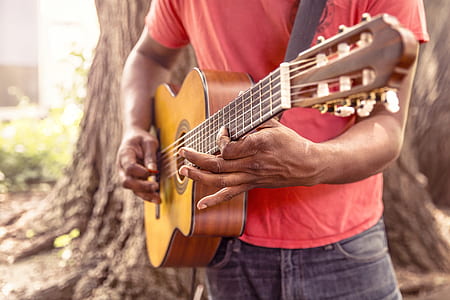 man wearing red t-shirt playing brown classical acoustic guitar near tree