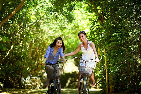 two girl's riding on bicycle passing through green tunnel of trees