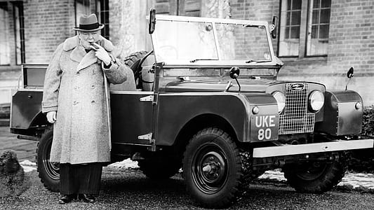 grayscale photo of man in front of vehicle wearing button-up coat