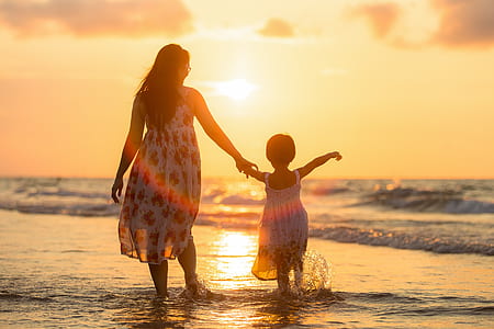 woman wearing white and red sleeveless dress walking with girl on sea shore during sunset
