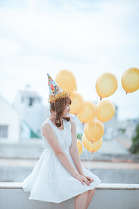 brown haired woman in white sleeveless dress holding yellow balloons
