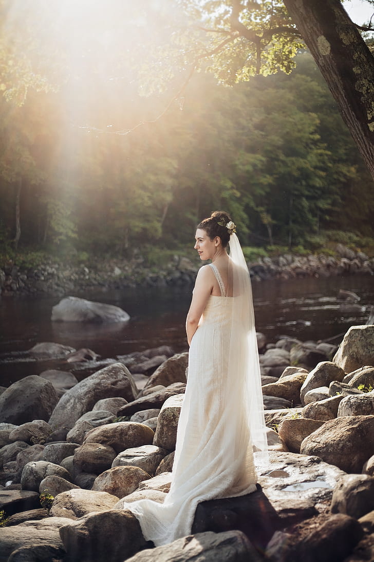 woman wearing white sleeveless wedding gown standing on rocks near body of water