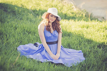 woman wearing blue and white sleeveless polka-dot circle dress and white sun hat sitting on grass field