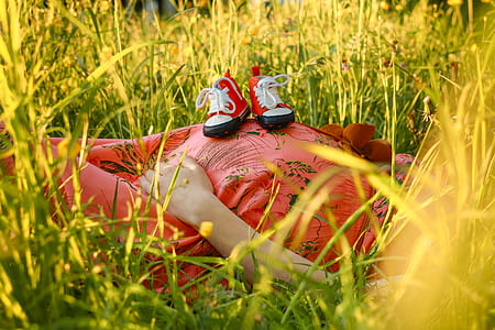 woman wearing red dress lying on grass with toddlers sneakers on her belly
