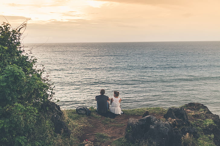 man and woman sitting on cliff near wave of sea