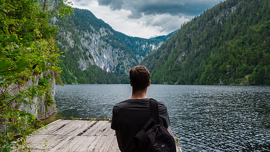 man in black shirt standing on dock beside lake surrounded by mountains