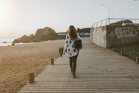 woman in white and black star print long-sleeved shirt and black denim jeans walking on brown wooden pathway near body seashore