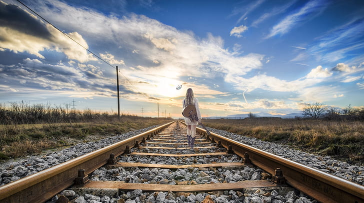 woman wearing white dress holding guitar walking on brown steel train rail road