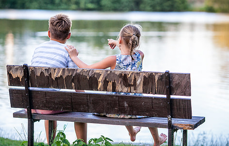 girl and boy sitting on brown wooden bench near body of water at daytime