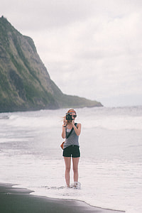 woman holding black instant camera standing on beach during daytime