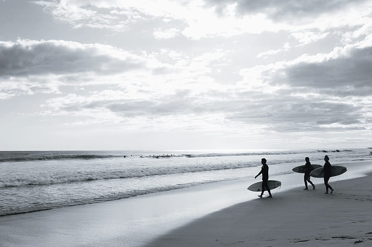 grayscale photo of three people holding surfboards walking on seashore